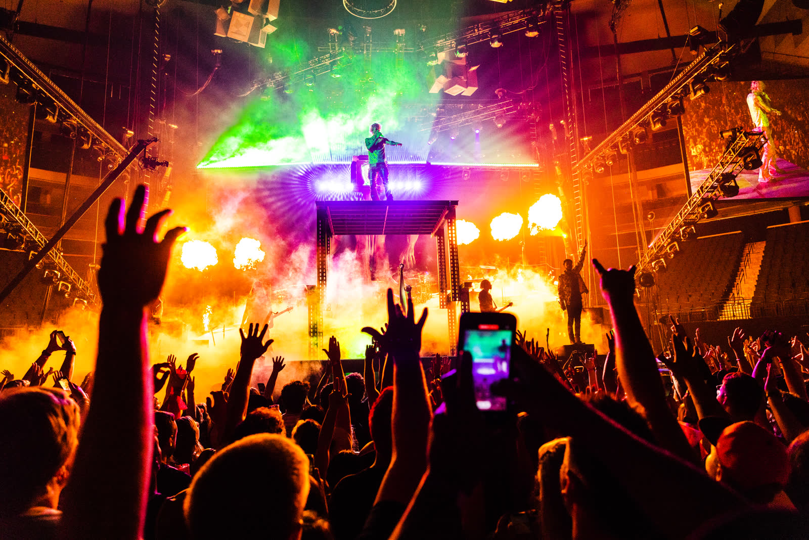 Shot from inside the crowd: Machine Gun Kelly on a raised platform under green light as flame pillars fire, phones and horns raised, Mainstream Sellout tour.
